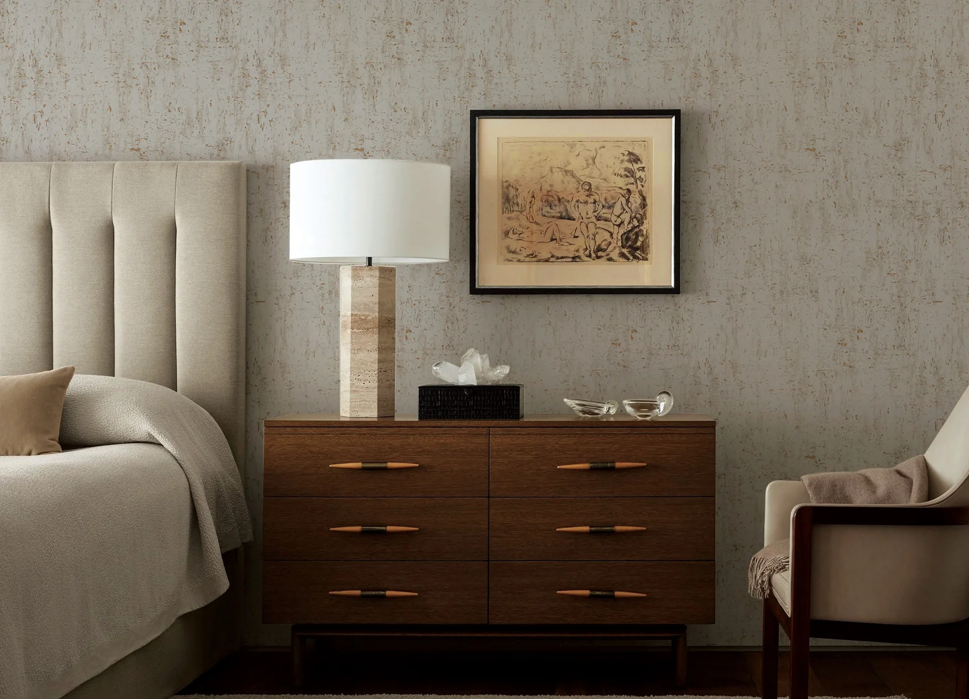 Neutral bedroom with a softly textured wallcovering behind a walnut chest of drawers, travertine lamp and framed print